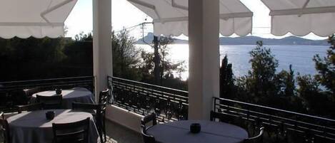 An outdoor terrace with round tables and chairs, white canopies, and a view of a body of water with mountains in the background.