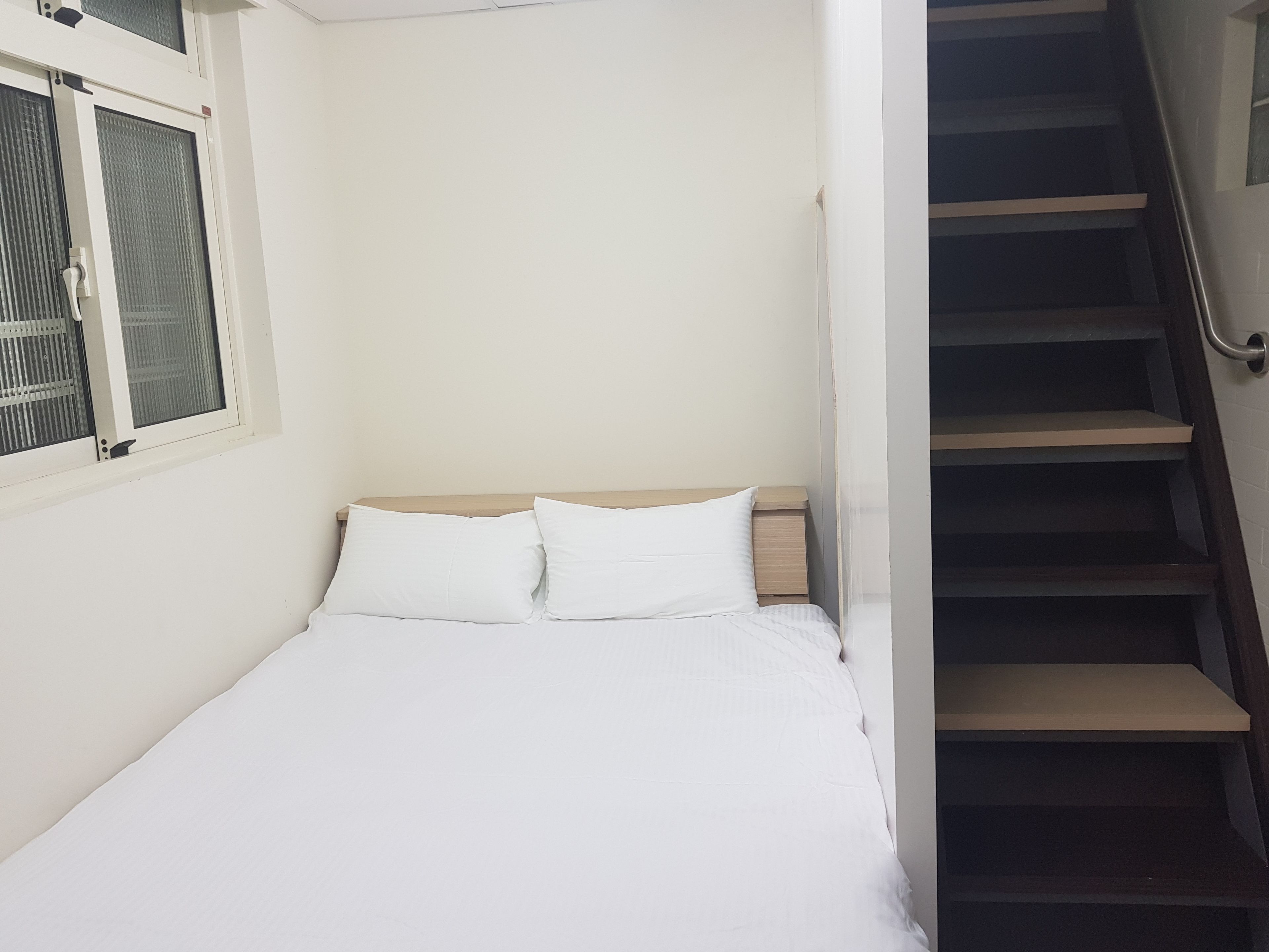 Uma cama pequena e bem arrumada, com roupa de cama branca, num quarto com paredes brancas e uma janela com persianas.