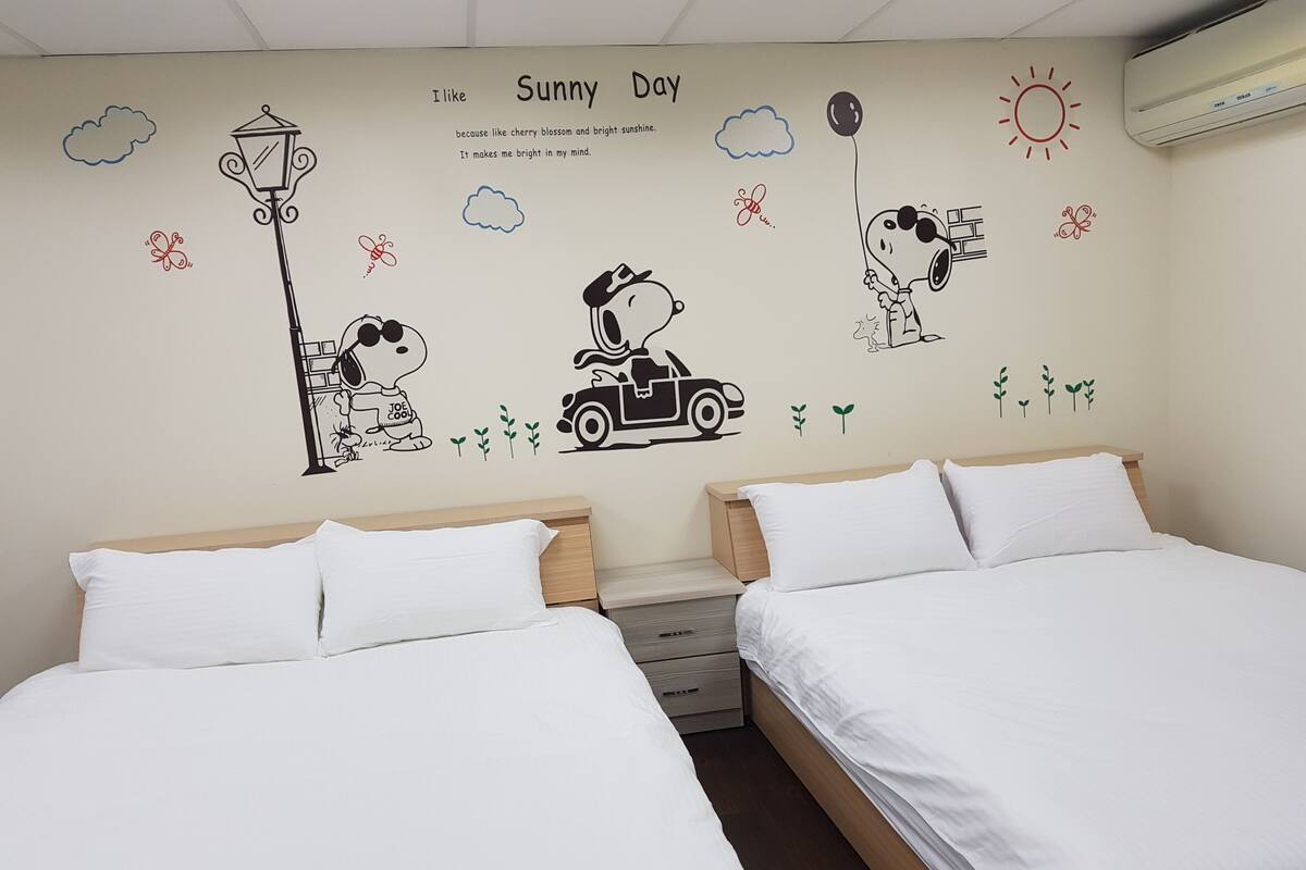 Une chambre d’hôtel avec deux lits, une fresque murale représentant Snoopy, un lampadaire et une voiture.