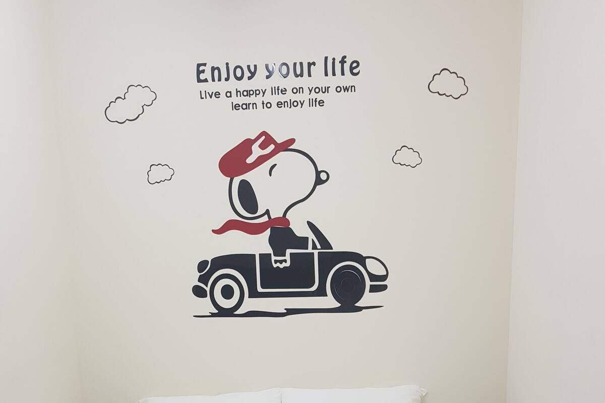 Une chambre à coucher avec un lit, un autocollant mural représentant Snoopy au volant d’une voiture, et un texte motivant.
