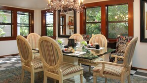 A dining area with a glass table surrounded by striped chairs, a chandelier, and large windows with views of greenery. - The Giraffe Inn at Old Parks Manor (Napa)
