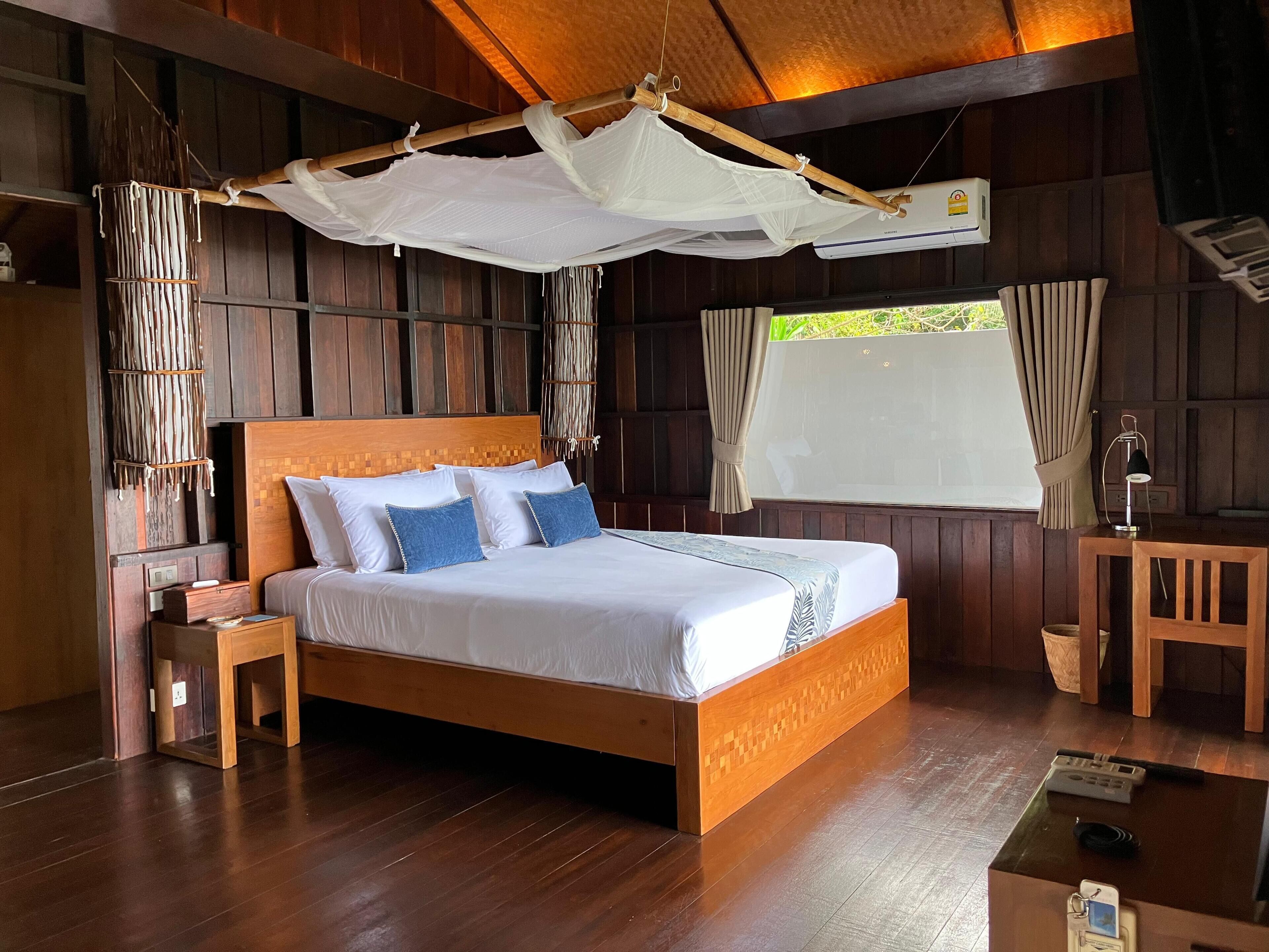 A wooden-framed bed with white bedding and blue pillows, a bedside table, a hanging mosquito net, and a window with curtains.
