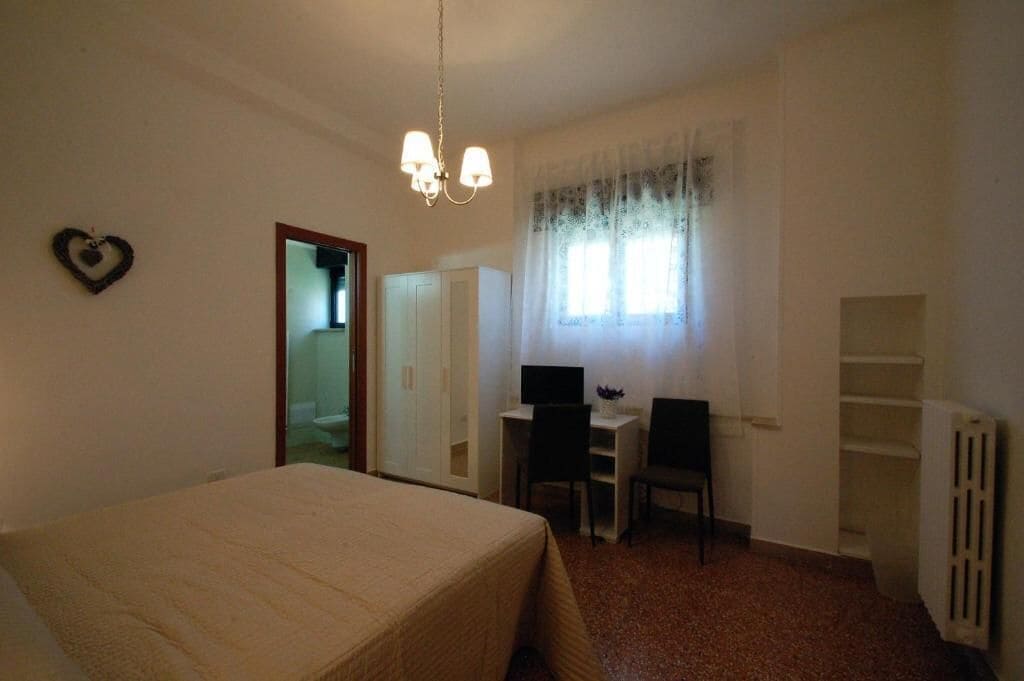 Una camera da letto con un letto, una scrivania, due sedie, un computer, un termosifone e una finestra con tende.