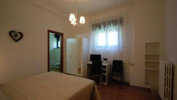 A bedroom with a bed, a desk, two chairs, a computer, a radiator, and a window with curtains.