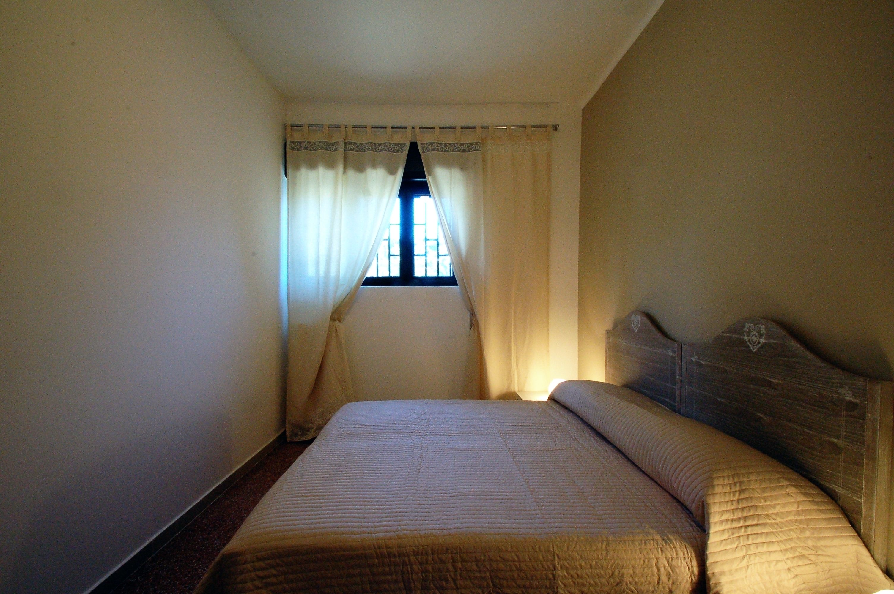 Una habitación pequeña y estrecha con una cama individual, una ventana con cortinas y un cabecero de madera.