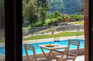 Una mesa de madera con dos sillas junto a una piscina, un plato de fruta y dos copas de vino tinto.