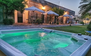 Une piscine à l’eau légèrement verdâtre, entourée d’espaces de détente en plein air avec des parasols, d’un mur en pierre et d’une pelouse bien entretenue.