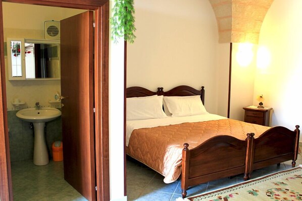 Une chambre à coucher avec un lit en bois, une table de chevet, une commode, une lampe fixée au mur, et une salle de bain avec un lavabo et un miroir.