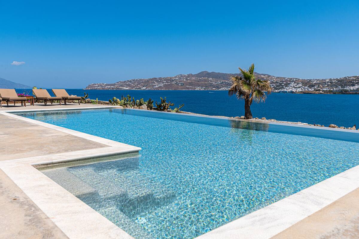A clear blue pool with lounge chairs and a palm tree, overlooking a coastal landscape.