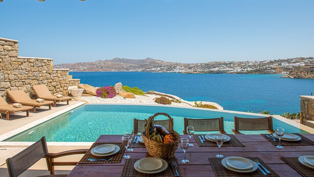 Ein Essbereich am Pool mit einem Tisch für zwei Personen, mit Blick auf das klare blaue Meer und eine gebirgige Landschaft.