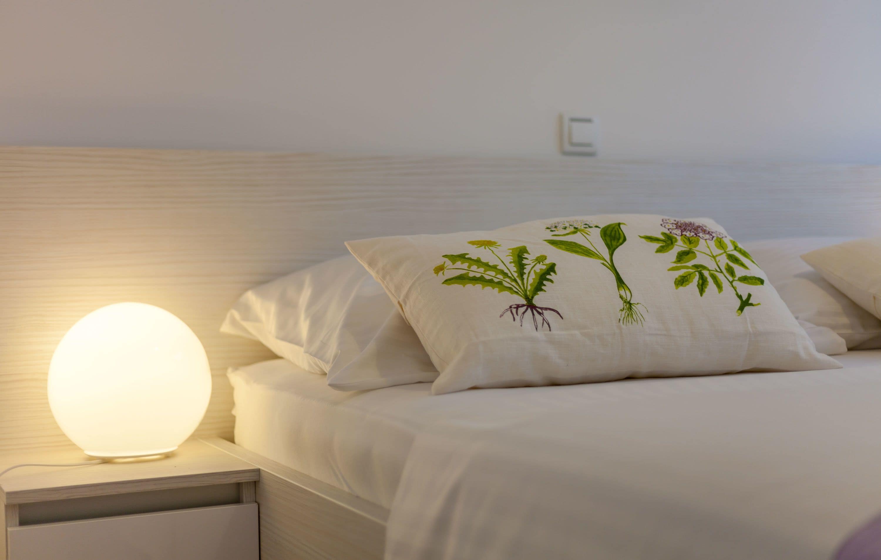 A neatly made bed with white bedding and pillows, a round bedside lamp, and a wall-mounted light switch.