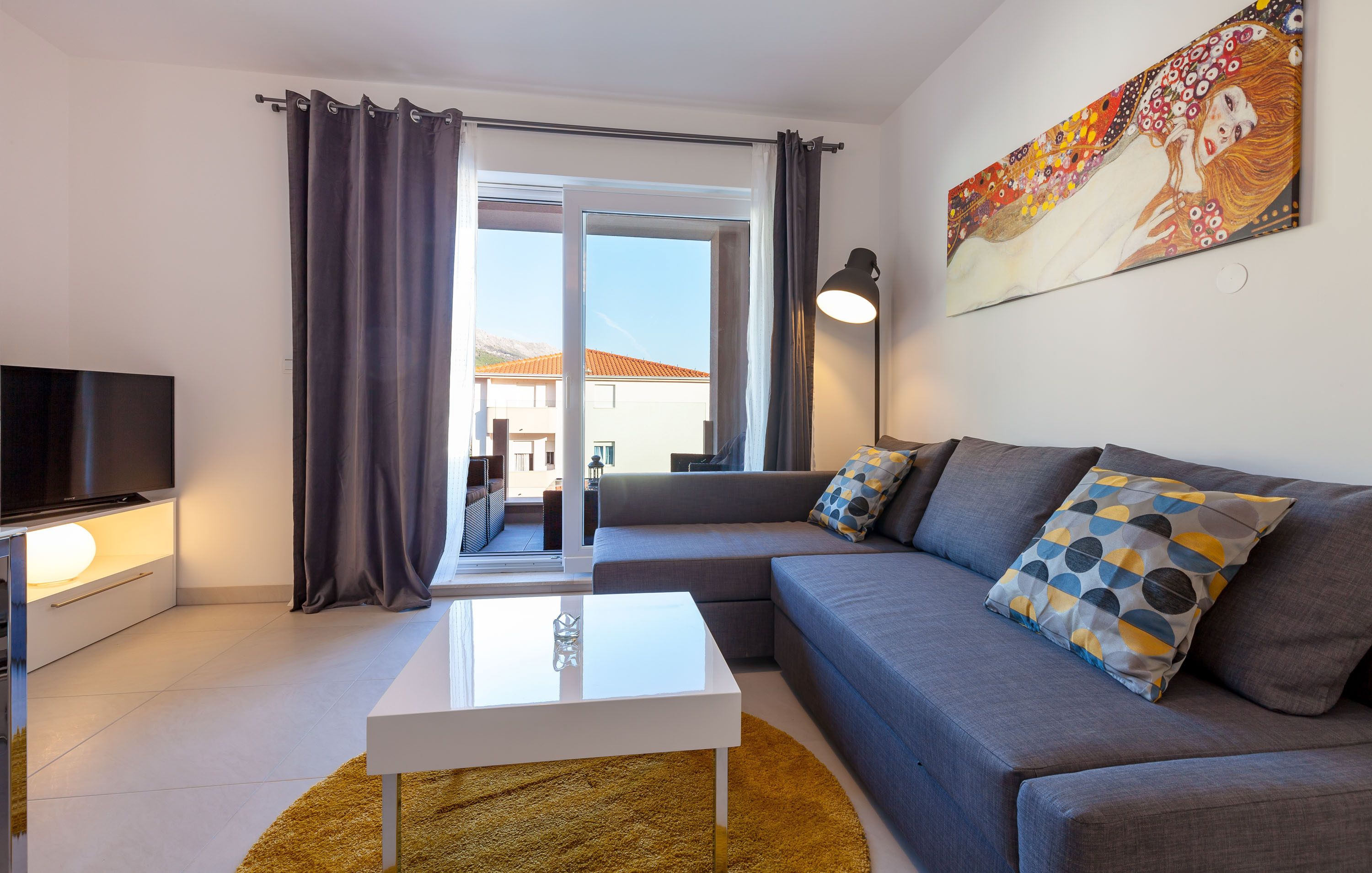 A modern living room with a flat-screen TV, a white coffee table, a grey sofa with patterned cushions, and a large window with curtains.