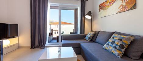 A modern living room with a flat-screen TV, a white coffee table, a grey sofa with patterned cushions, and a large window with curtains.