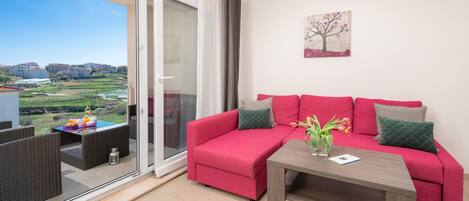 A red sofa with green cushions, a wooden coffee table, and a view of a green landscape through a glass door.