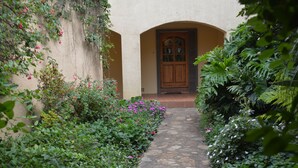 A stone pathway leading to a wooden door with a glass panel, surrounded by lush greenery and flowering plants.