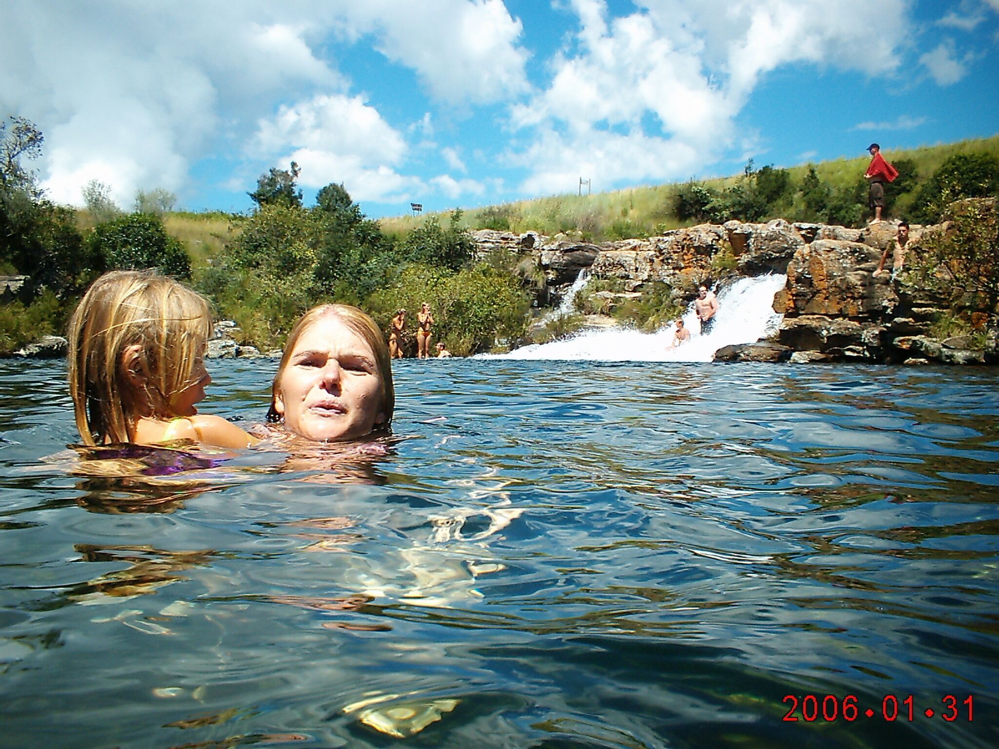 A natural hot spring with clear water, surrounded by greenery and a small waterfall.