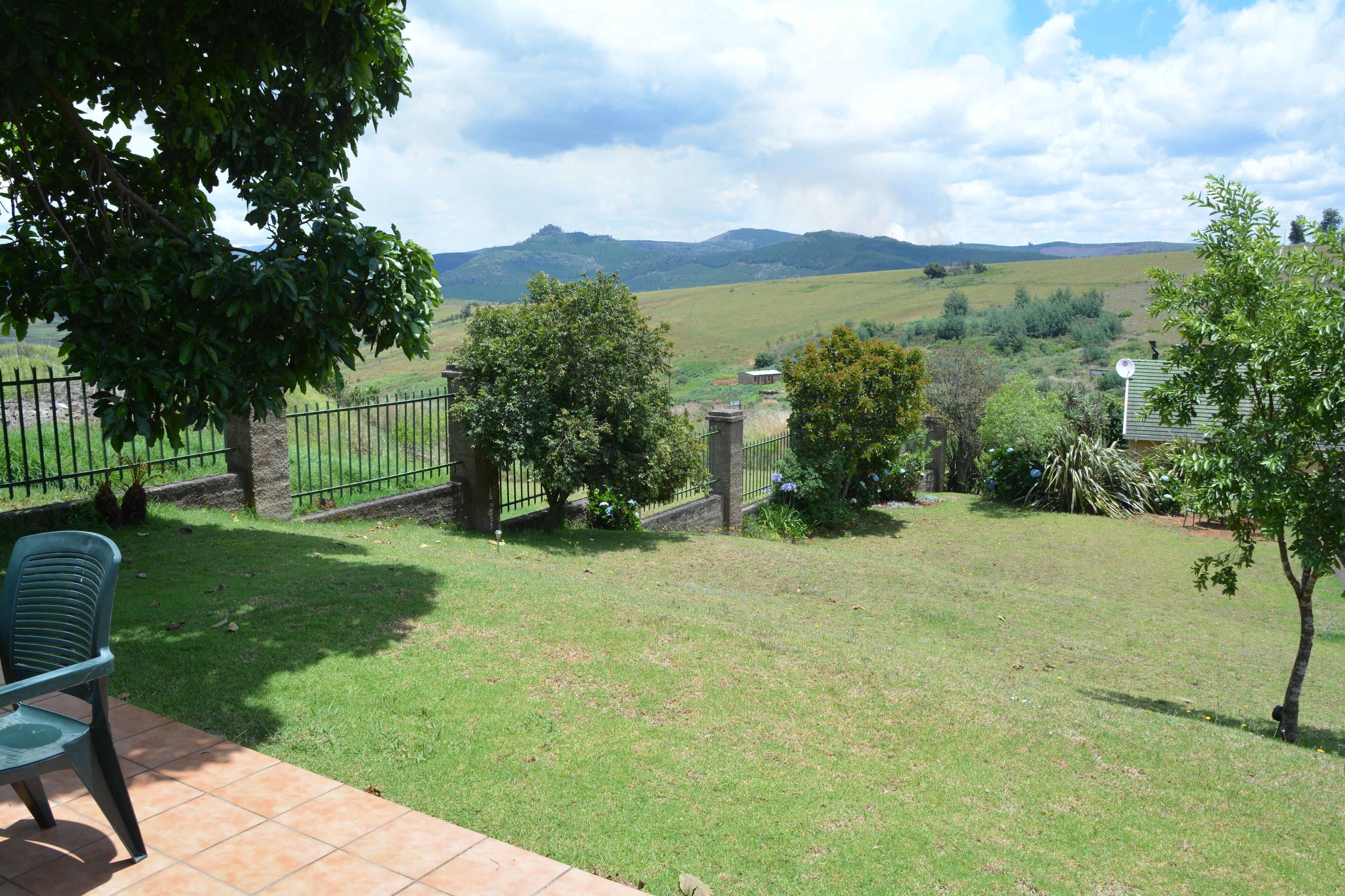 Un patio con una silla verde, un muro de piedra y vistas a un campo cubierto de césped con árboles y una montaña al fondo.