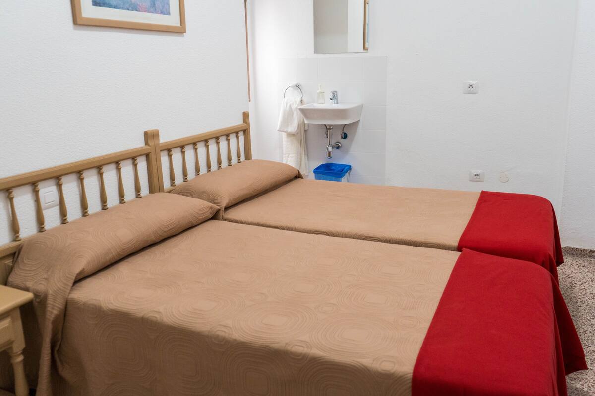 Una habitación con dos camas, cabecera de madera, lavamanos blanco y cubo de basura azul.