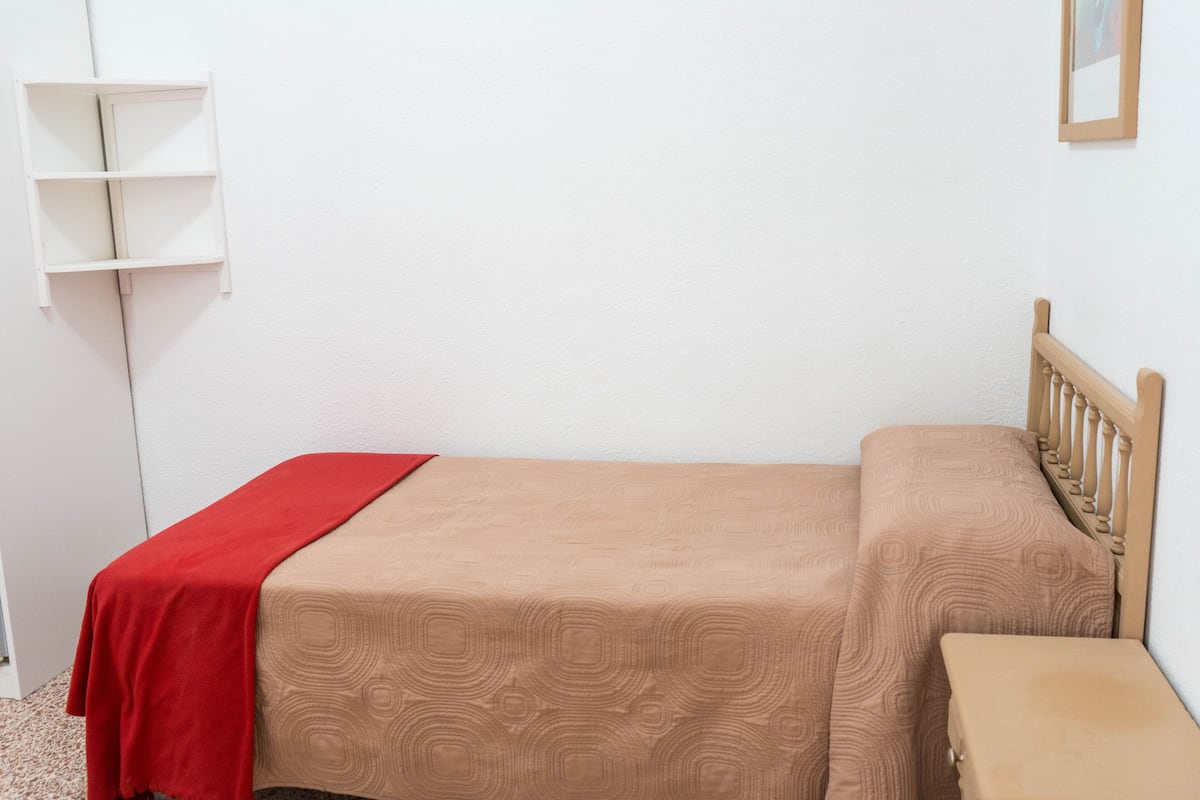 Una cama individual con cabecera de madera, una colcha roja y un cubrecolchón color beige.