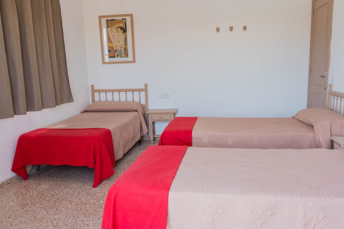Una habitación con dos camas individuales, cada una con cubrecama rojo y beige, una mesita de noche de madera, una pequeña mesa y un cuadro en la pared.