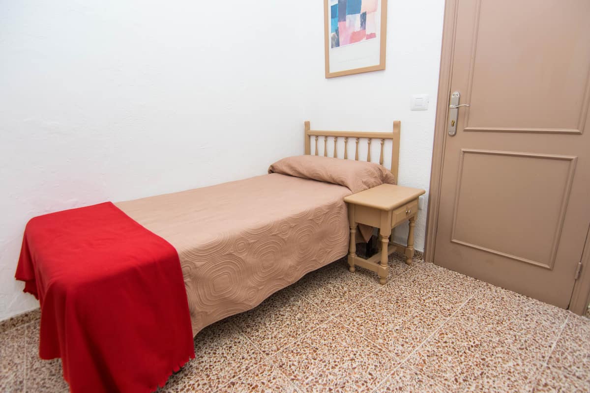 Una cama individual con una colcha roja, una mesita de noche de madera y una puerta.