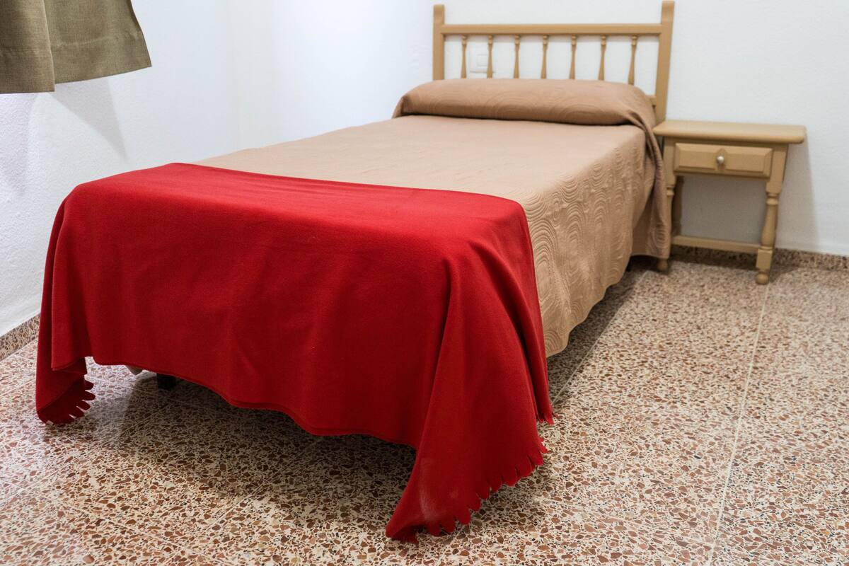 Una cama con colcha roja y cabecera de madera, una mesita de noche de madera con un cajón, y un suelo moteado.