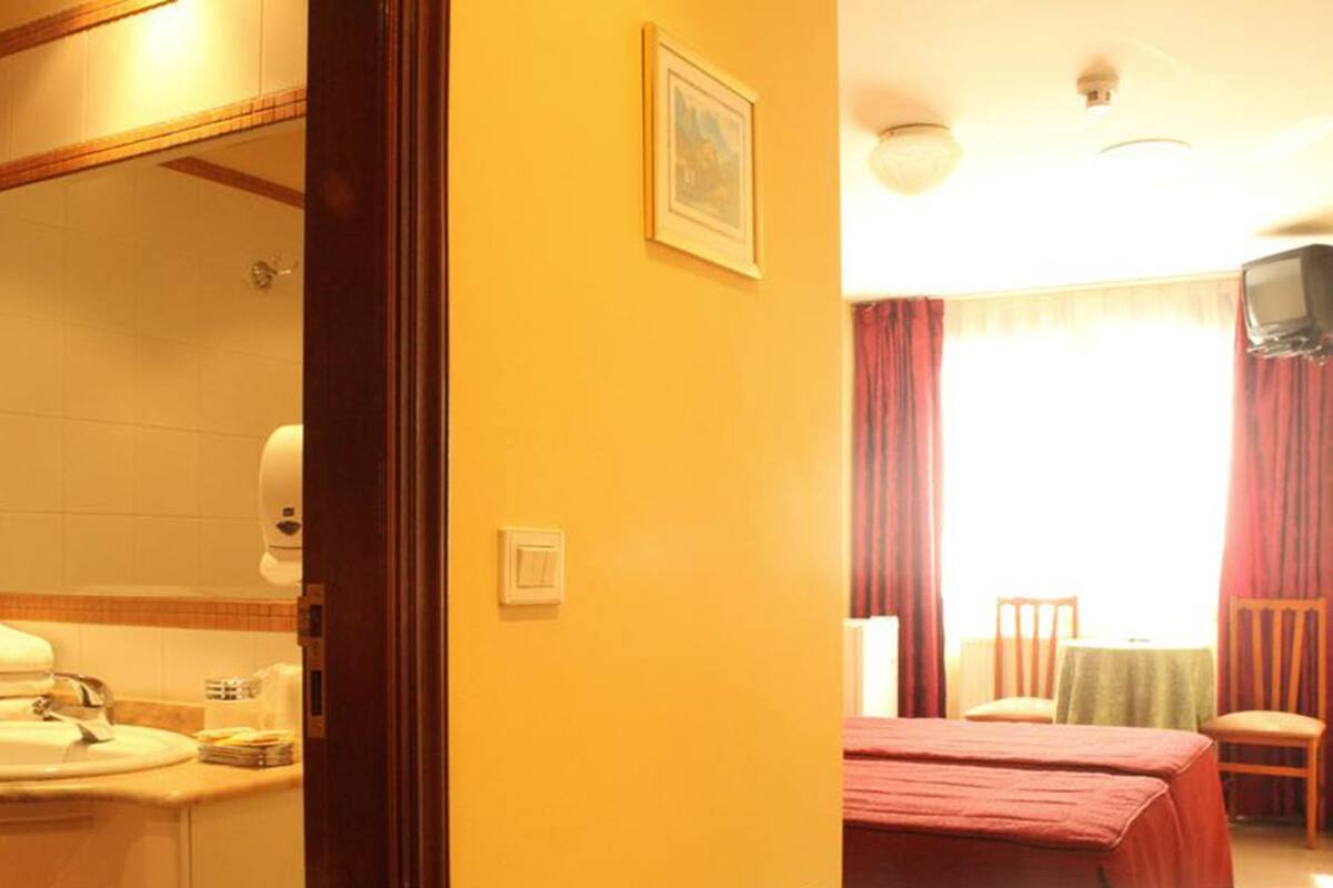 Un baño de hotel con lavabo, espejo y retrete. Hay una cama con colcha roja y un televisor en la habitación.