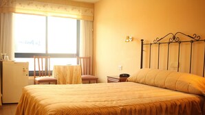 Une chambre d’hôtel comprenant un lit, une petite table, une chaise, un bureau, un réfrigérateur et une fenêtre avec des rideaux.