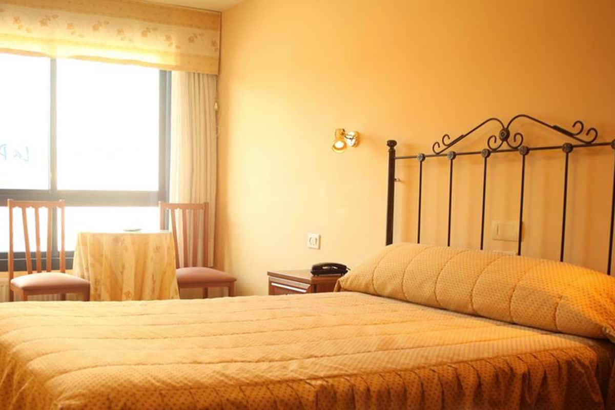 Habitación de hotel con cama, una mesita, una silla, escritorio, refrigerador y ventana con cortinas.
