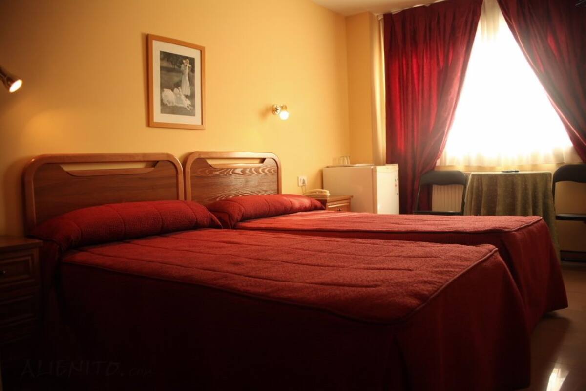 Habitación de hotel con dos camas, una mesita de noche, una lámpara, un cuadro en la pared y una ventana con cortinas.