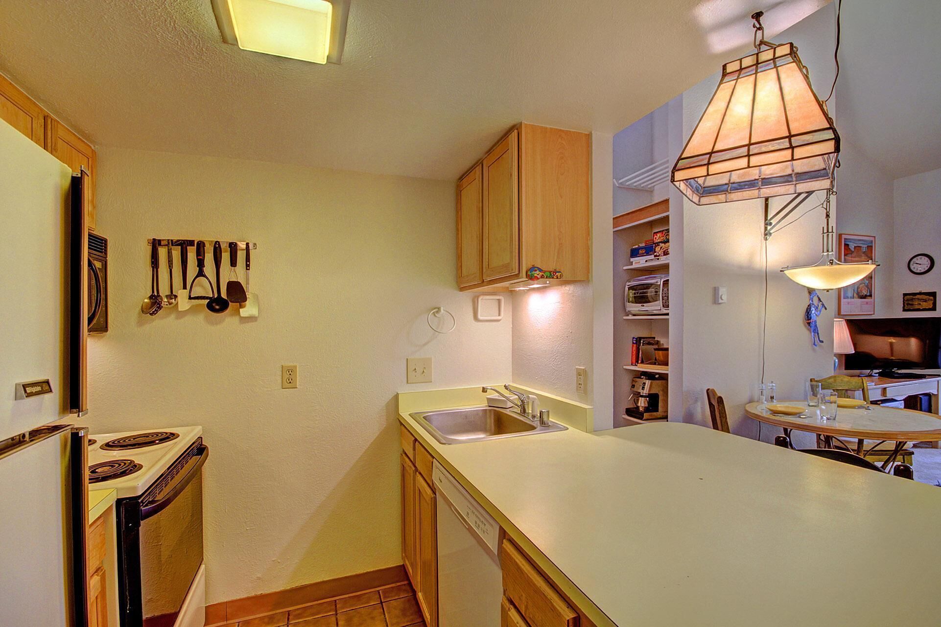 A compact kitchen with wooden cabinets, a built-in dishwasher, a stove, and a hanging light fixture.