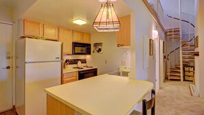 Una cocina compacta con armarios de madera, un refrigerador blanco, un microondas y una estufa. Hay una pequeña isla con una silla, una escalera que sube a un altillo, y una lámpara colgante sobre la isla.
