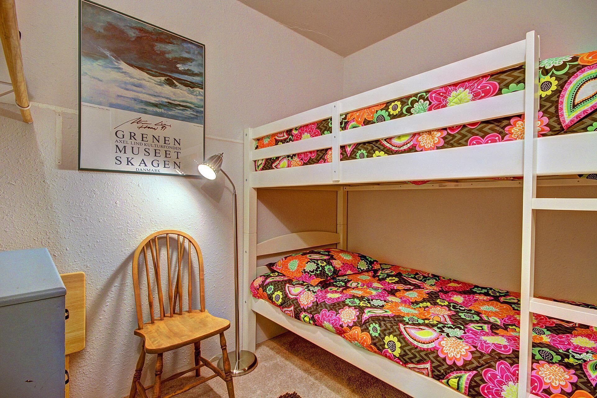 Una cama nido con ropa de cama floral de colores, un escritorio con una silla y un cuadro en la pared.