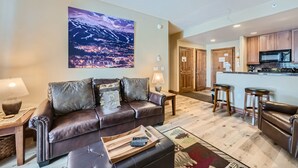 A modern living room with a brown leather sofa, a wooden coffee table, and a cityscape painting on the wall. - Highland Greens Lodge 210 1 Bedroom Condo by RedAwning (Breckenridge)