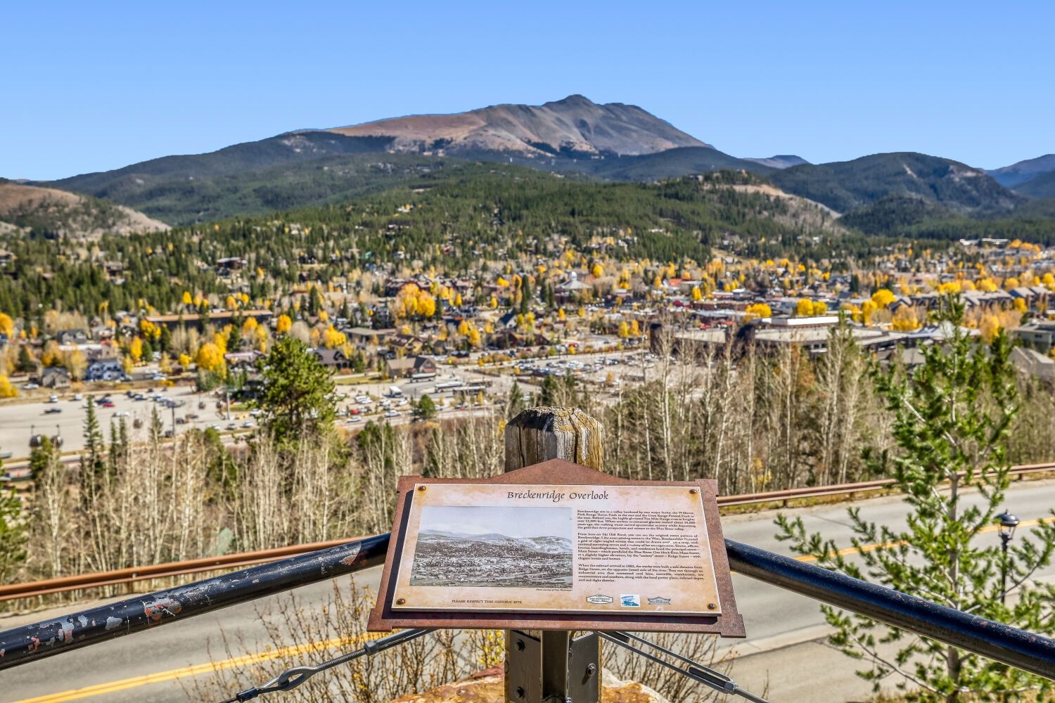 A scenic overlook with a signboard, a road, and a view of a town with autumn-colored trees and mountains in the background.