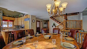 A dining area with a wooden table set for six, surrounded by leather chairs. A chandelier hangs above, and a staircase with wooden railings is visible in the background.