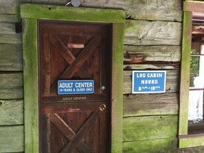 A wooden door with a sign indicating an adult center for 18 years and older, and a log cabin hours sign from 8 AM to 10 PM. - 57sw - Fireplace - Wifi -  Sleeps 6 2 Bedroom Home by RedAwning (Deming)