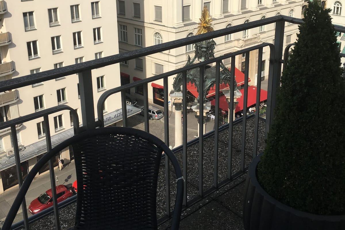 Un balcón con una silla de mimbre y una planta en maceta, con vista a una calle con edificios y un coche rojo.