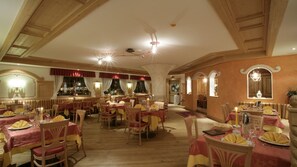 A spacious dining area with multiple tables set for dining, wooden beams on the ceiling, and a chandelier. - Hotel Al Polo (Ziano di Fiemme)