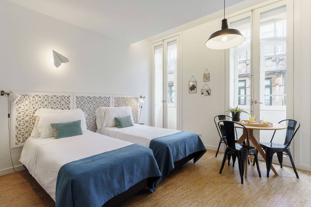 A hotel room with two beds, a dining table, chairs, and a window with a view of buildings.