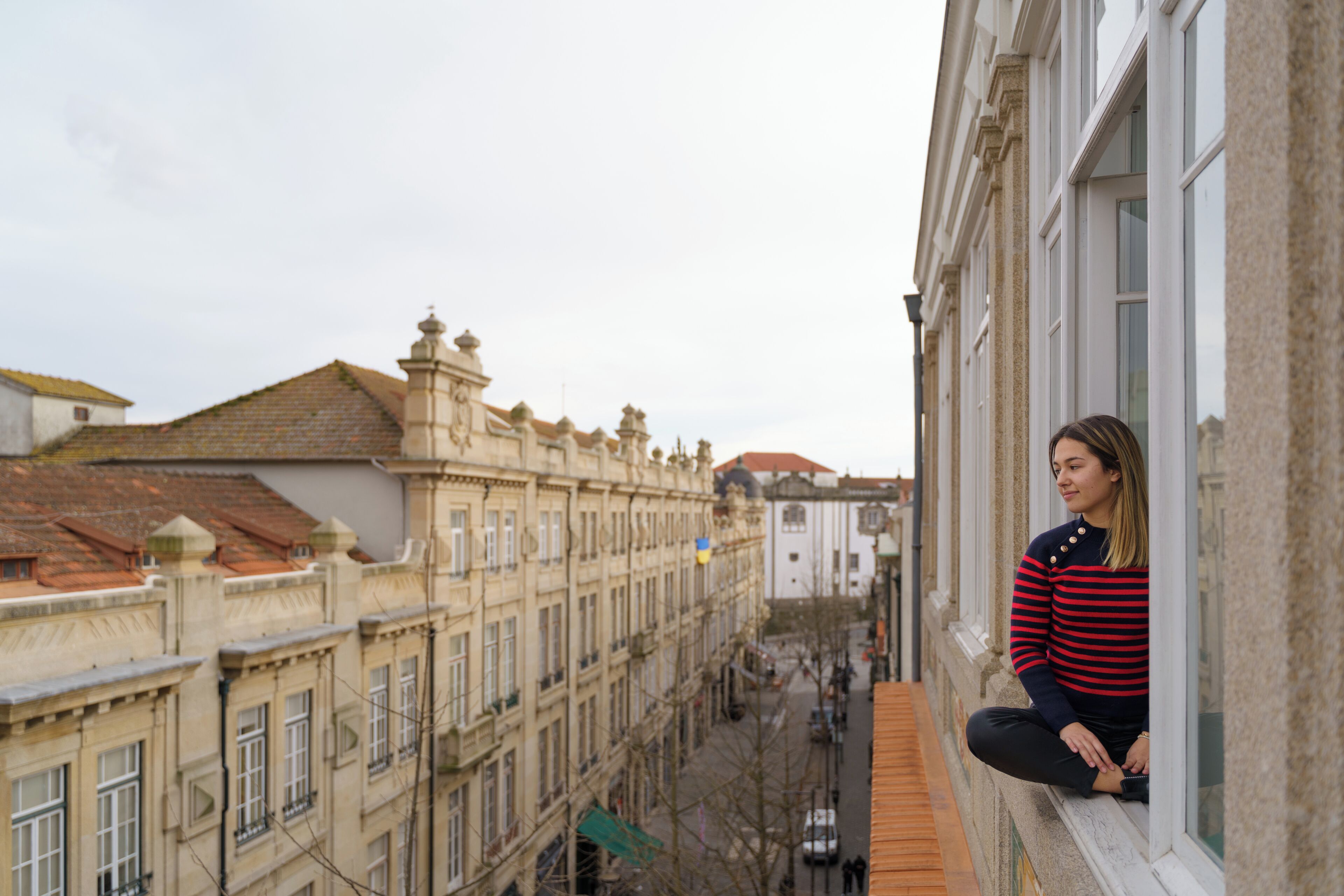 A person sitting on a balcony with a view of a cityscape, including buildings with classical architecture and a clear sky.