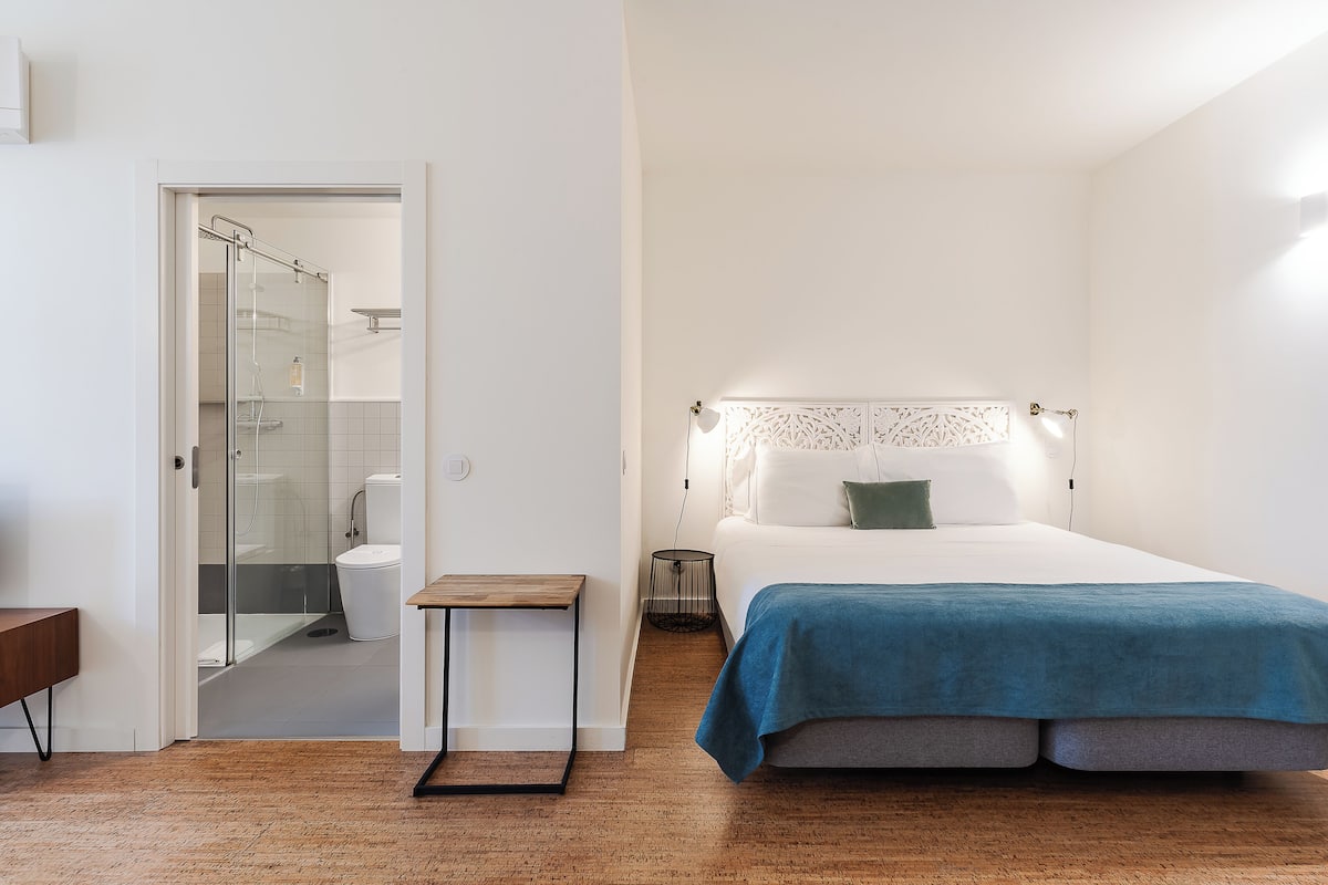A bedroom with a bed, bedside table, and a small table. There is a bathroom with a toilet and a shower.