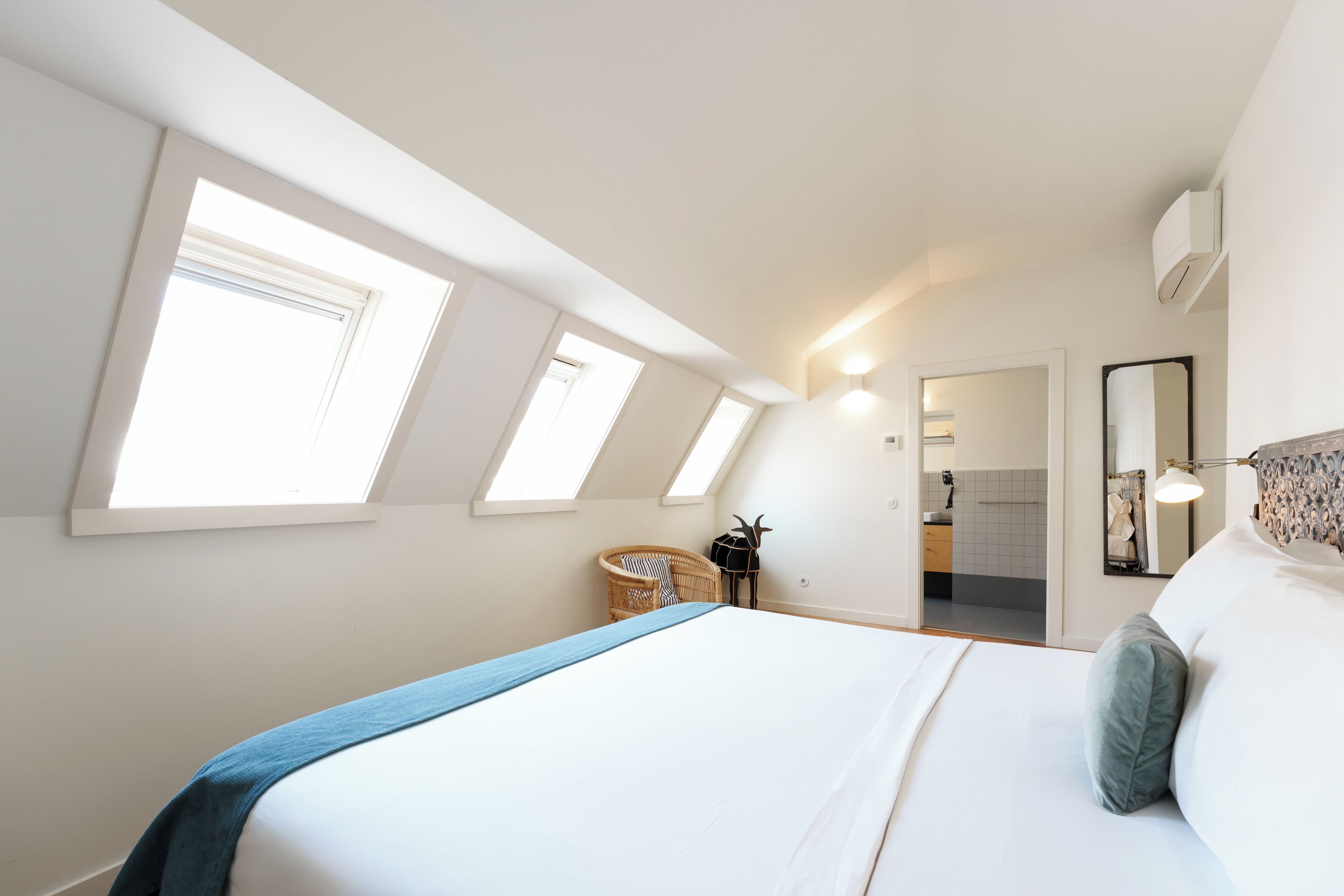 Een slaapkamer met een groot bed, dakramen, een rieten stoel, een klein tafeltje en een spiegel aan de muur.
