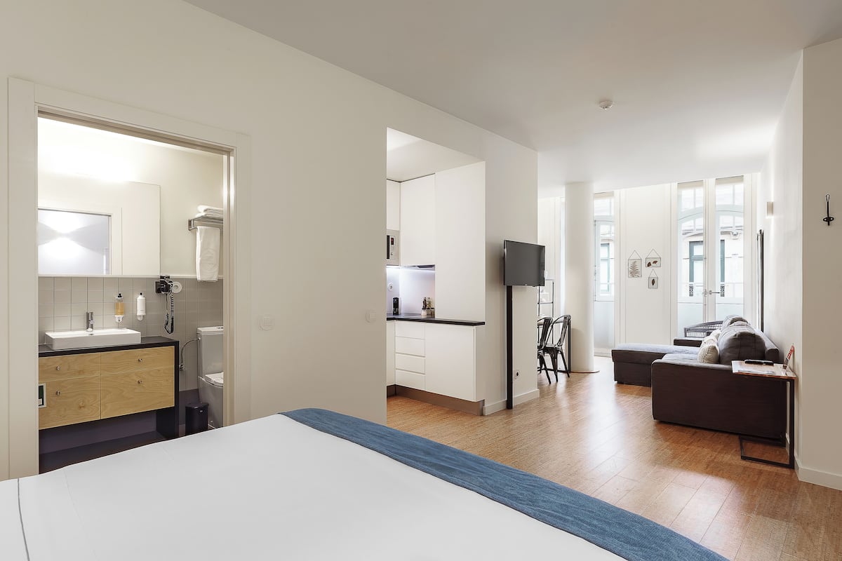 A modern hotel room with a bed, a bathroom, a kitchenette, and a living area with a sofa and TV.