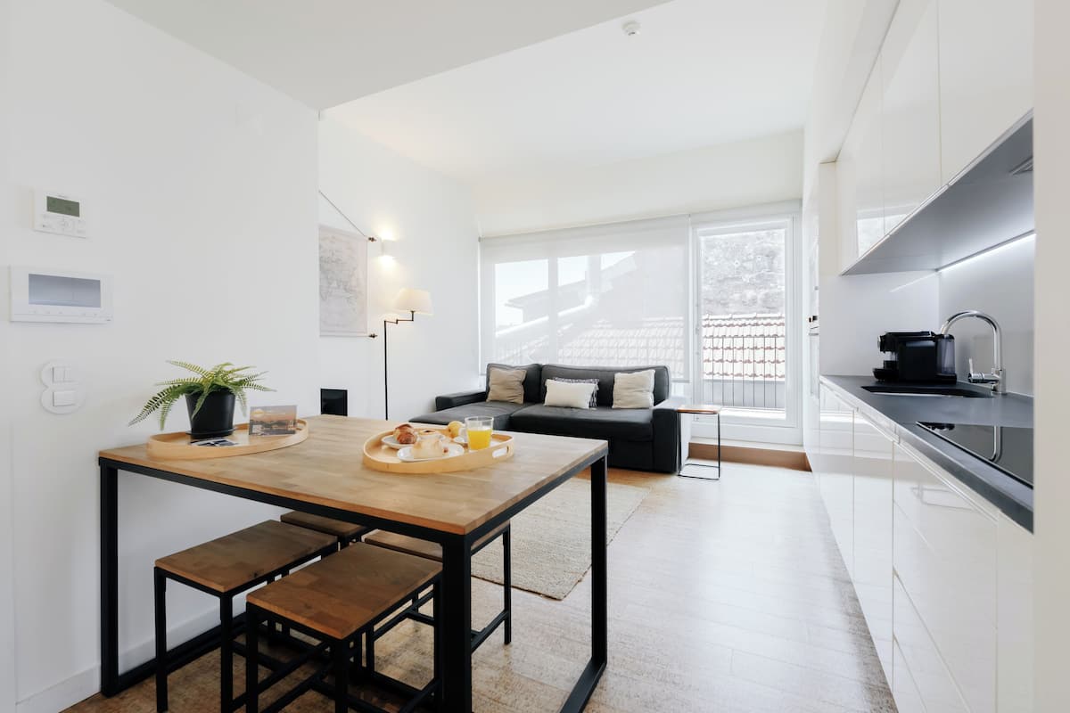 A modern kitchen with a dining area, a sofa, and a large window.