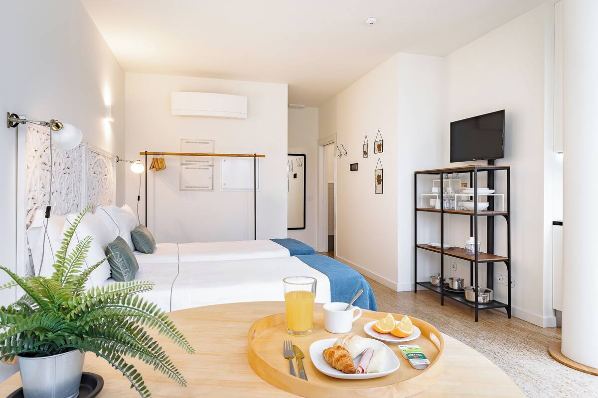 A modern hotel room with a bed, a dining table with breakfast, a TV, and a plant.