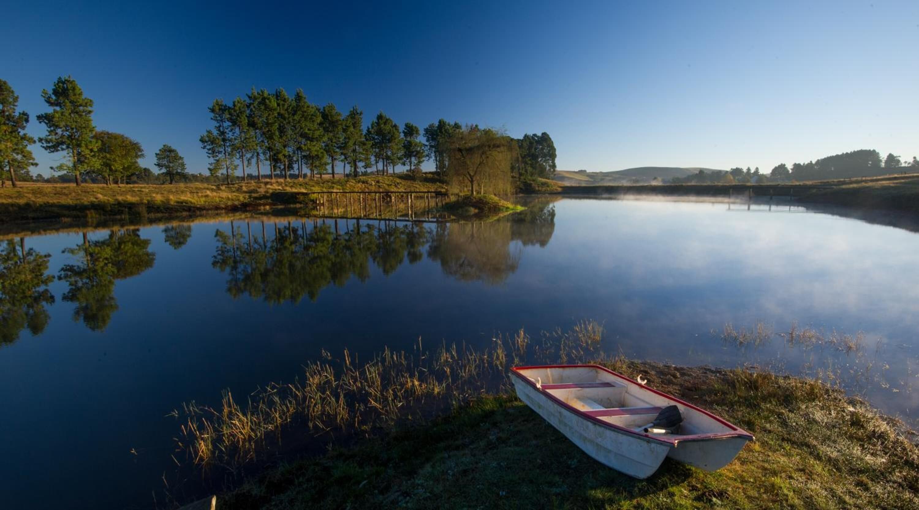A calm lake with a small boat on the shore and a wooden fence in the background.