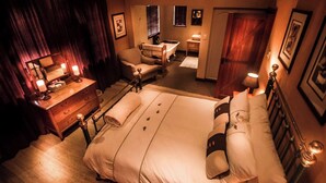 Ein Hotelzimmer mit einem großen Bett, einem Schreibtisch mit Lampe, einem Sessel, einem kleinen Tisch ebenfalls mit Lampe und einem durch eine offene Tür einsehbaren Badezimmer.