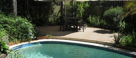 Una piscina con borde de azulejos, rodeada por una terraza con mesa y sillas, y una sombrilla blanca que da sombra.
