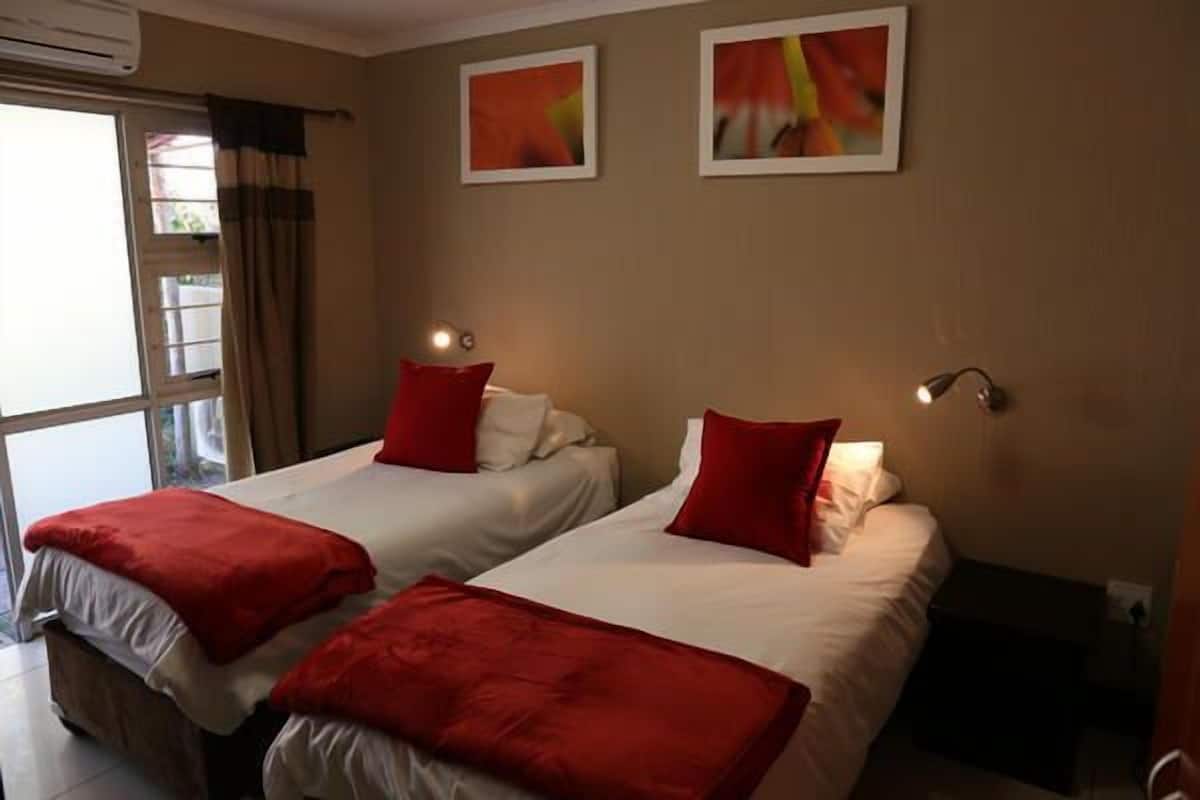 Habitación de hotel con dos camas, cada una con ropa de cama roja y almohadas blancas, una ventana con cortinas y dos lámparas fijadas a la pared.